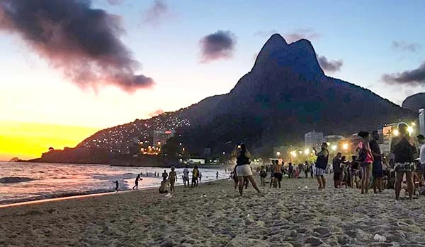 Ao pôr do sol na praia com a paisagem icônica do Morro Dois Irmãos do Rio de Janeiro ao fundo, pessoas desfrutam do final de tarde na orla