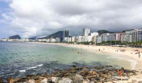 Praia de Copacabana no Rio de Janeiro com vista para o mar, areia branca, ondas, edifícios altos ao fundo e céu nublado.