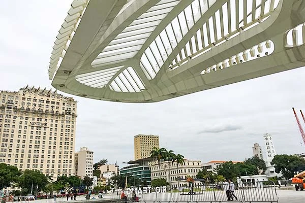 Imagem de um edifício moderno com uma estrutura arquitetônica incomum, localizado em um ambiente urbano com prédios clássicos e árvores, sob céu nublado.