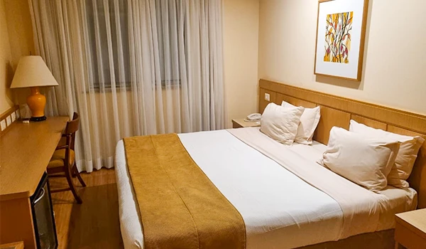 Quarto de hotel aconchegante com cama de casal, roupa de cama branca, almofadas, decoração moderna, painel de madeira, cortinas de tecido e iluminação suave.