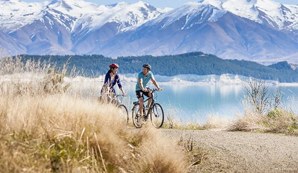 Foto de um casal de bicicleta em uma paisagem de campo, com lago e montanhas nevadas na Lua de Mel na Nova Zelândia.