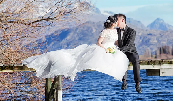Foto de um casal de noivos sentados em um pier rústico de madeira, tendo ao fundo montanhas e mar na Lua de Mel na Nova Zelândia.