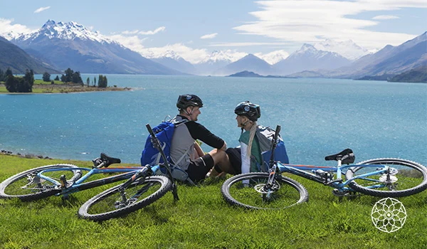 Foto de um casal com bicicletas na grama, sentados com linda vista da água e das montanhas.