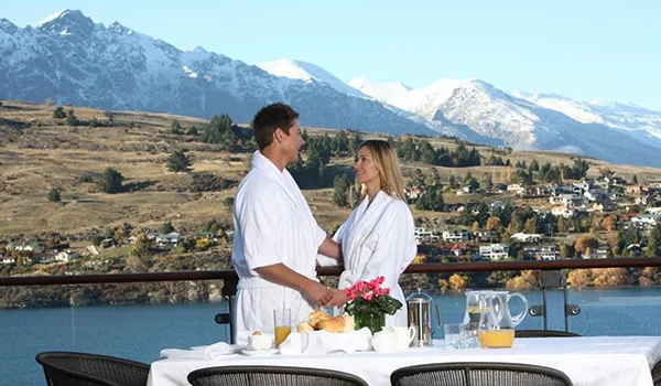 Foto de um casal jovem em Lua de Mel na Nova Zelândia na varanda de um hotel com mesa preparada para o café da manhã, tendo uma vista bonita da região.