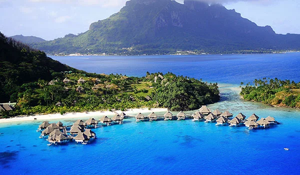 Fotografia aérea de uma lagoa azul-turquesa de Bora Bora, com bangalôs sobre a água, uma ilha verde ao centro e uma montanha imponente ao fundo.