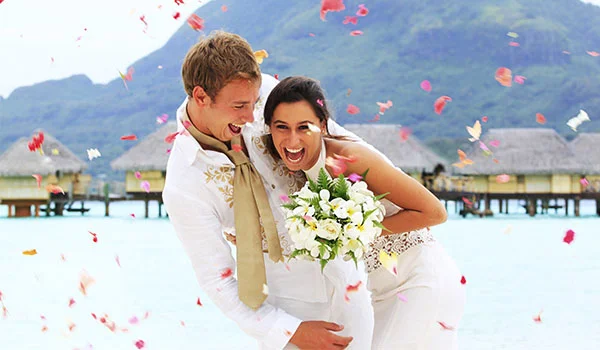 Casal recém-casado sorrindo e abraçado à beira de uma lagoa de águas cristalinas, com uma paisagem montanhosa ao fundo. A noiva segura um buquê de flores brancas e o casal veste roupas claras. Pétalas coloridas são lançadas ao ar ao redor deles.
