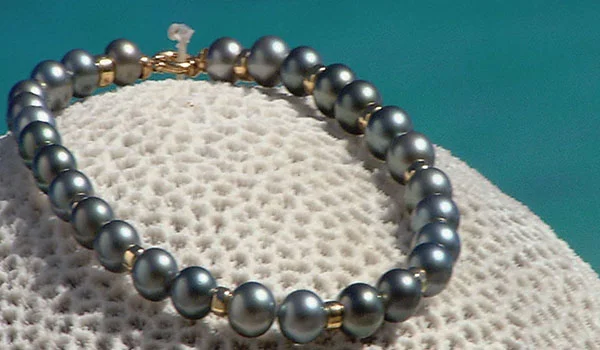 Colar de pérolas negras e prateadas sobre uma superfície branca, com fecho dourado na parte superior.
