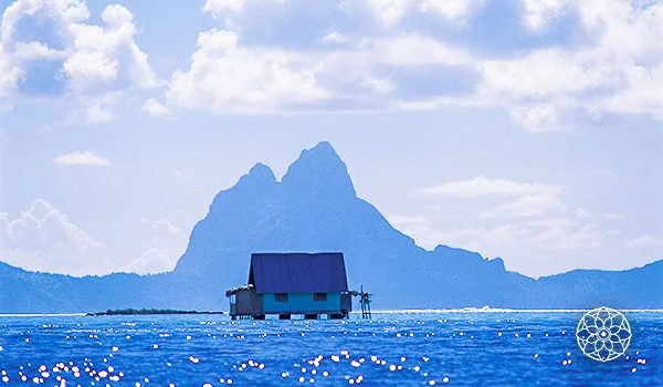 Lagoa azul-turquesa com uma pequena cabana sobre palafitas no centro, tendo ao fundo uma imponente montanha vulcânica recortada contra um céu azul com nuvens brancas. A água brilhante reflete a luz do sol, criando pequenos pontos luminosos na superfície.
