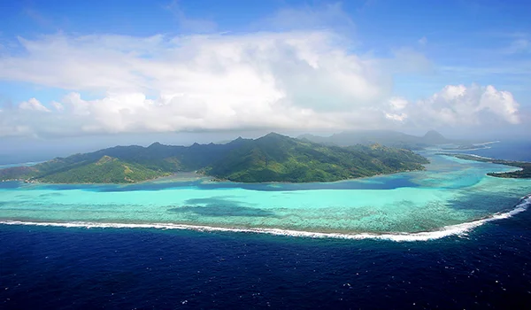 Ilha tropical vista do alto, cercada por águas cristalinas azul-turquesa e um recife de coral, com vegetação verde exuberante, montanhas ao centro, céu azul com nuvens brancas e o oceano azul-escuro em primeiro plano.