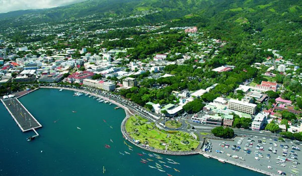 Vista aérea de Papeete, no Taiti, mostrando a área urbana junto ao mar, com uma marina, ruas, estacionamentos e edifícios cercados por vegetação tropical e montanhas ao fundo.