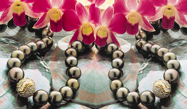 Colar de grandes pérolas negras com detalhes dourados nas extremidades, disposto sobre uma superfície espelhada, acompanhado de várias flores de orquídea rosa e branca.