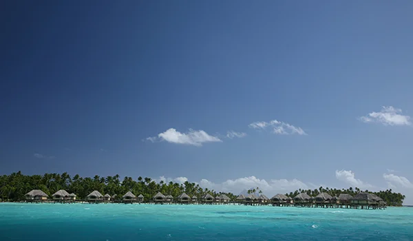 Ilha tropical com águas cristalinas azul-turquesa, céu azul com poucas nuvens e uma fileira de bangalôs sobre a água ao longo da costa, cercados por vegetação verdejante.