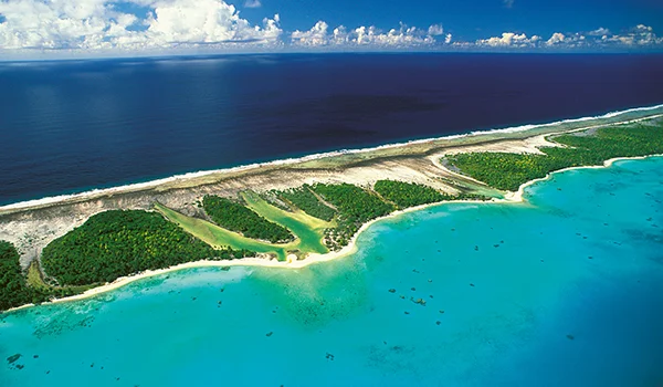 Vista aérea de um atol tropical, com águas azul-turquesa rasas, recifes de coral, pequenas áreas de vegetação verde e uma longa faixa de praia branca separando a lagoa do oceano azul profundo. Nuvens brancas aparecem no horizonte.
