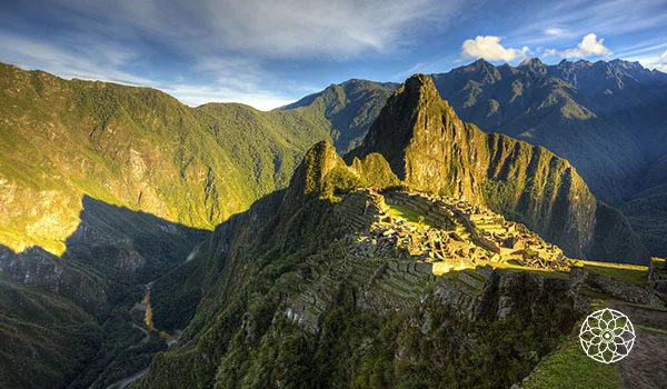 Vista panorâmica de Machu Picchu, com as antigas ruínas incas sobre um estreito terraço entre montanhas verdes e íngremes. Ao fundo, destacam-se picos cobertos de vegetação e algumas nuvens, iluminados pela luz do sol.