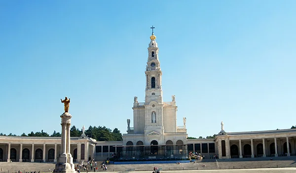 Santuário religioso de arquitetura branca, com uma alta torre central encimada por uma cruz e ladeado por longas galerias com colunas e arcos. Em frente, há uma coluna com uma estátua dourada no topo e várias pessoas na praça, sob um céu azul claro.