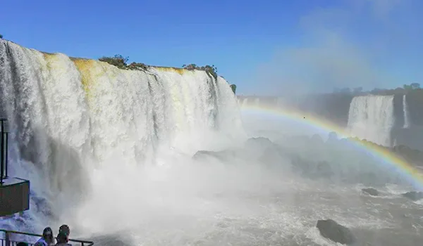 Grandes quedas-d’água despencam de paredões rochosos, formando uma intensa nuvem de névoa sobre o rio. Um arco-íris aparece sobre a água entre as cataratas, sob um céu azul claro.