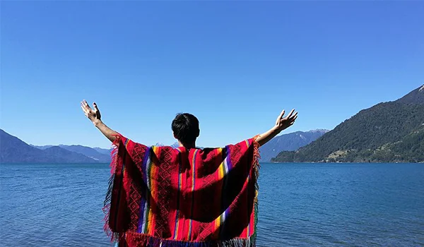 Pessoa vista de costas, com os braços erguidos, usando um poncho colorido com padrões geométricos, diante de um grande lago azul cercado por montanhas. O céu está limpo e azul, criando uma paisagem ampla e ensolarada.