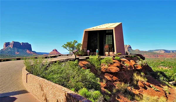 Edificação moderna de formato angular, construída sobre uma formação rochosa, com grandes janelas voltadas para uma paisagem de montanhas avermelhadas. Ao redor, há vegetação típica de região árida e uma área pavimentada, sob um céu azul intenso.