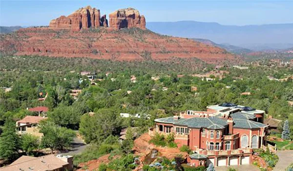 Vista panorâmica de Sedona, no Arizona, com uma formação rochosa de arenito vermelho ao fundo, cercada por vegetação verde. Em primeiro plano, há casas e edifícios espalhados pela paisagem, sob um céu azul claro.