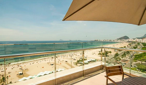 Vista de uma varanda de um hotel com guarda-sol, vista panorâmica de uma praia de areia, mar e horizonte ao fundo, com céu claro e algumas nuvens, ideal para relaxar e aproveitar o sol.
