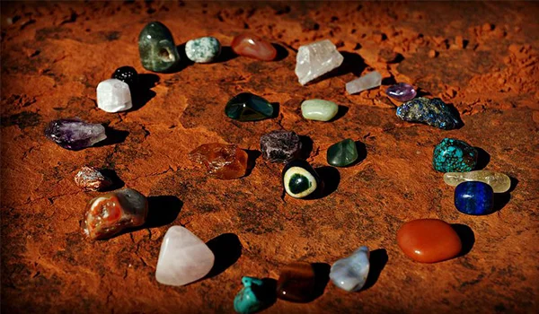 Diversas pedras e cristais polidos de diferentes cores, formatos e tamanhos estão espalhados sobre uma superfície de pedra marrom-avermelhada. Há exemplares verdes, azuis, roxos, brancos, alaranjados e translúcidos, iluminados por uma luz intensa.