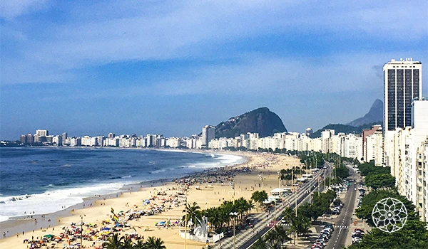 Vista panorâmica da Praia de Copacabana no Rio de Janeiro, com alto movimento de turistas, linhas de ônibus e edifícios residenciais ao fundo, incluindo o Pão de Açúcar ao longe.