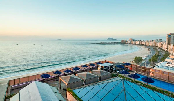 Vista de uma praia com mar tranquilo ao pôr do sol, apartamentos e cabanas na orla, cidade ao fundo, ideal para férias e lazer na praia.