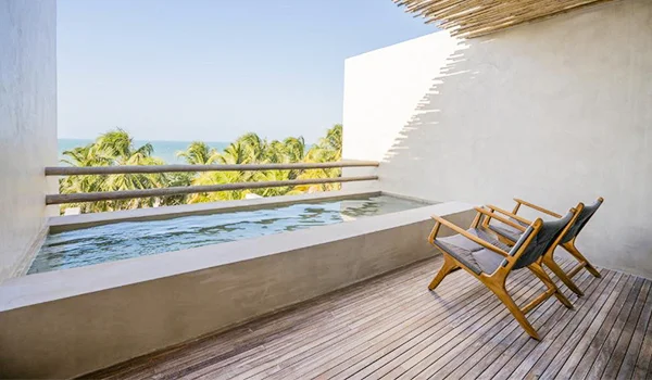 Varanda de hotel com piscina privativa, deck de madeira e duas cadeiras de descanso, com vista para palmeiras e o mar ao fundo.