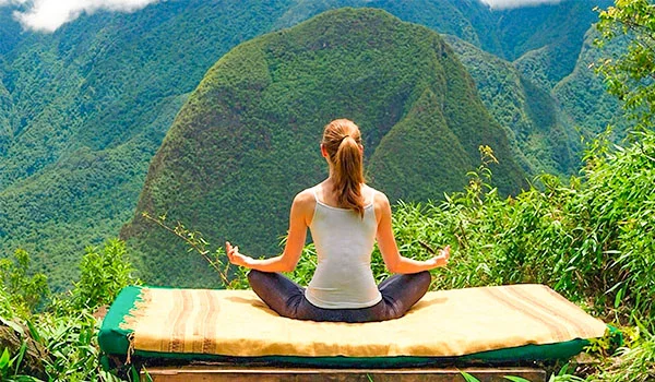 Mulher sentada em posição de lótus sobre um tapete, meditando diante de uma paisagem montanhosa exuberante e coberta de vegetação. Ao fundo, grandes montanhas verdes envoltas por névoa; ao redor, folhagens tropicais.