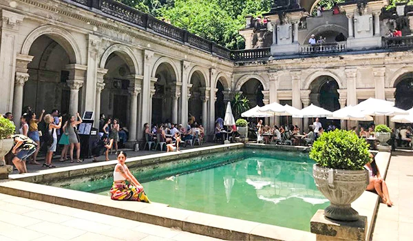 Vista do pátio com piscina, cadeiras de descanso, pessoas aproveitando o ambiente ao ar livre, cercado por arquitetura clássica e áreas verdes.