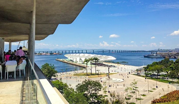 Vista panorâmica de uma área de lazer com espaço aberto, árvores, pessoas e um amplo calçadão, sob um céu azul com algumas nuvens.