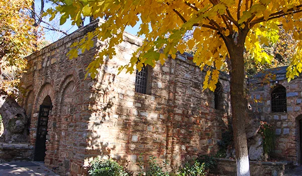 Edificação histórica de pedra e tijolos, com pequenas janelas gradeadas e uma porta em arco, parcialmente coberta pela sombra de árvores. As folhas amarelas e alaranjadas indicam uma paisagem de outono.