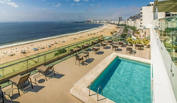 Vista de uma piscina na cobertura de um hotel de luxo com vista para a praia e o mar, em uma cidade movimentada, com cadeiras de descanso e uma fachada moderna.