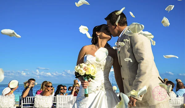 Noivos se beijam durante uma cerimônia de casamento ao ar livre, diante do mar. A noiva segura um buquê de flores brancas, enquanto pétalas são lançadas ao redor do casal.