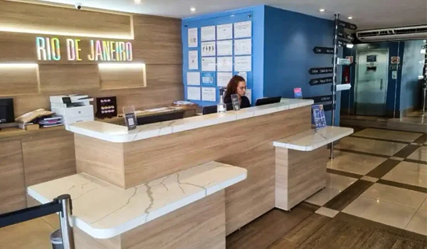 Recepção moderna de um hotel ou escritório em Rio de Janeiro, com design elegante, balcão de atendimento em madeira clara, equipe auxiliando clientes e sinalização colorida ao fundo.