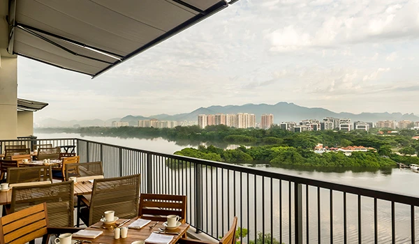 Vista de um espaço de lazer ao ar livre com mesas e cadeiras em um restaurante com vista para o rio, árvores e prédios na cidade ao fundo, sob céu parcialmente nublado. Cafeteria do Grand Hyatt, uma das atrações sobre o que fazer na Barra da Tijuca.