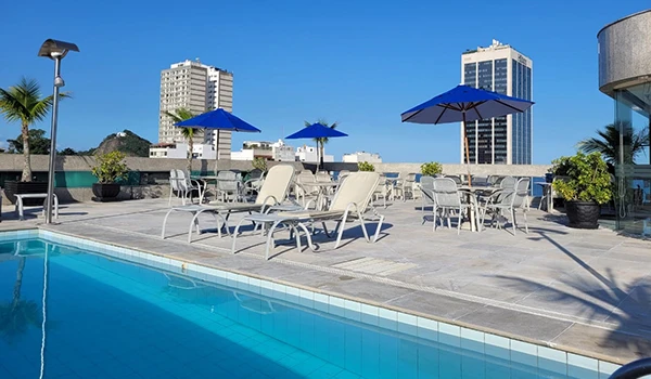 Área de lazer com piscina ao ar livre, espreguiçadeiras e guarda-sóis na cobertura de um prédio, com vista para edifícios altos ao fundo, ideal para relaxar e aproveitar o sol.