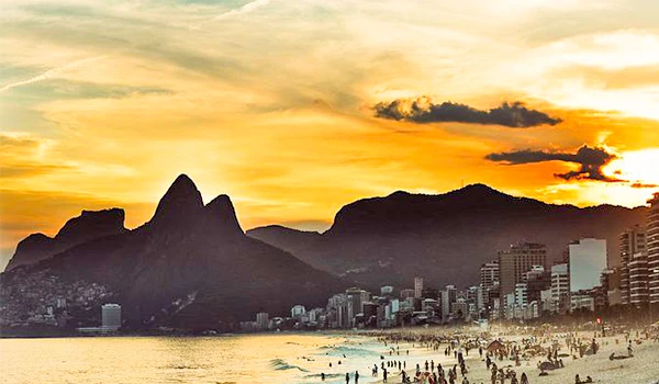 Praia no Rio de Janeiro ao entardecer com montanhas ao fundo e o céu colorido ao pôr do sol, destacando a beleza natural e urbana da cidade.