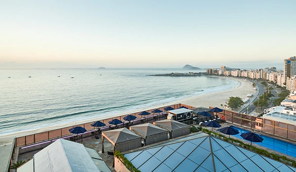 Vista panorâmica da orla de uma cidade costeira ao pôr do sol, com prédios, praia, espreguiçadeiras e cabanas na areia.
