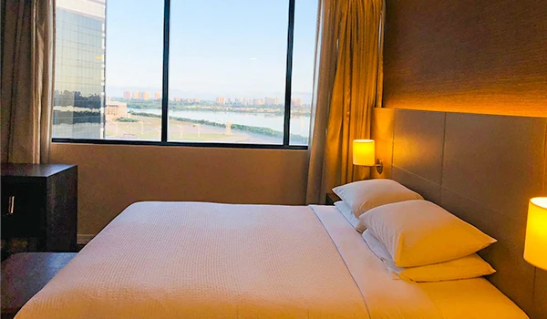 Quarto de hotel com cama de casal, roupas de cama brancas, iluminação suave e vista para a cidade através de uma grande janela. O que fazer na Barra da Tijuca.