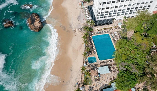 Vista aérea de uma piscina ao lado de uma praia de areia com ondas, coqueiros e edifícios ao fundo, criando um cenário de lazer e descanso à beira-mar.