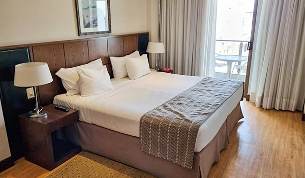 Quarto de hotel bem decorado com cama de casal, cortinas longas na janela e varanda com vista, ideal para hospedagem confortável.