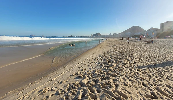 Praia de Copacabana no Rio de Janeiro com areia dourada, mar azul e horizonte com prédios e morros ao fundo.