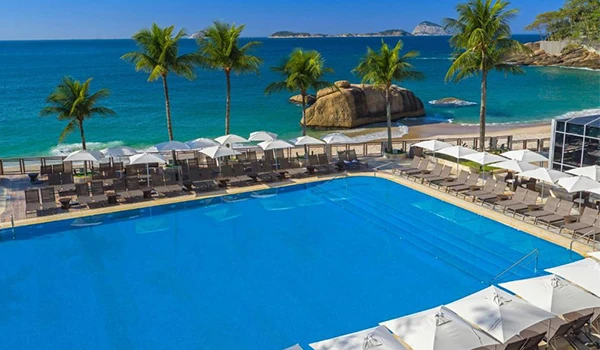 Piscina de resort à beira-mar com espreguiçadeiras e toldos brancos, palmeiras, mar azul e ilhas ao fundo, criando um cenário paradisíaco de férias.