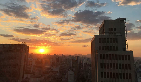 Vista de um pôr do sol em um horizonte urbano com edifícios altos, nuvens coloridas e o céu em tons de laranja e azul, criando uma paisagem visualmente impressionante.