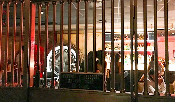 Imagem de um bar movimentado visto através das grades, com várias pessoas conversando e desfrutando de drinks em um ambiente acolhedor e moderno.