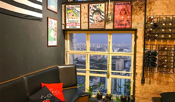 Interior de um espaço com vista urbana, decorado com quadros na parede, um sofá preto com almofadas e uma janela com vista para a cidade ao entardecer.