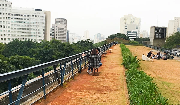 Imagem de uma pessoa sentada no terraço de um prédio com vista para uma cidade, plantas e pessoas ao fundo em um ambiente urbano com espaço aberto