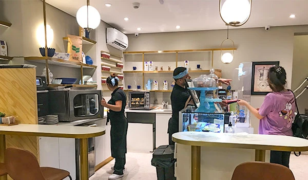Interior de uma cafeteria moderna com funcionários preparando bebidas e atendendo clientes, ambiente organizado e acolhedor.