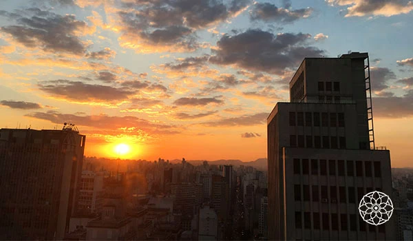 Tela de pôr do sol em uma cidade grande, com edifícios altos ao entardecer, nuvens coloridas no céu, criando uma cena urbana de fim de tarde.